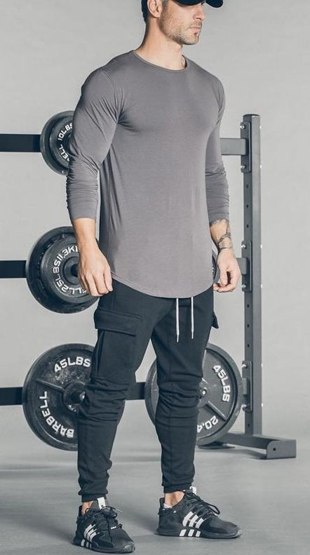 Man wearing Men's Cargo Joggers and long-sleeve shirt, posing in gym setting with weights in background.