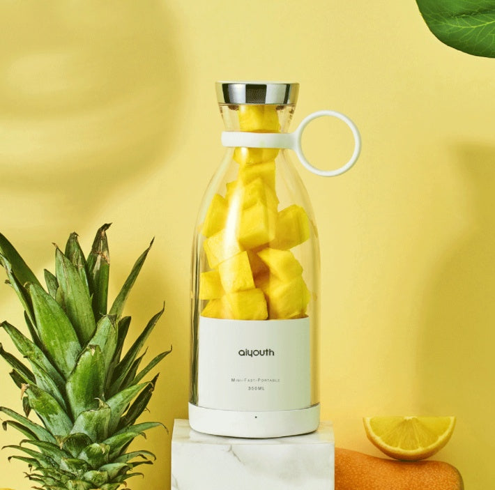 Portable electric mini juicer filled with fresh mango pieces, surrounded by pineapple and lemon, on a bright yellow background.