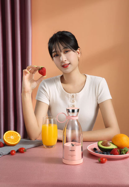 A woman holding a strawberry next to a portable electric mini juicer and a glass of orange juice on a table.